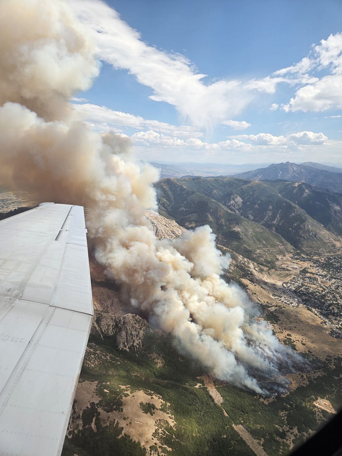 The view from inside an air attack plane circling a wildfire. The aircraft's white wing juts across the lower left of the frame, and below it a tall column of tan smoke boils up from fire burning along a steep mountain front, with a green valley and the edge of a town visible beyond and cumulus clouds in the blue sky above.