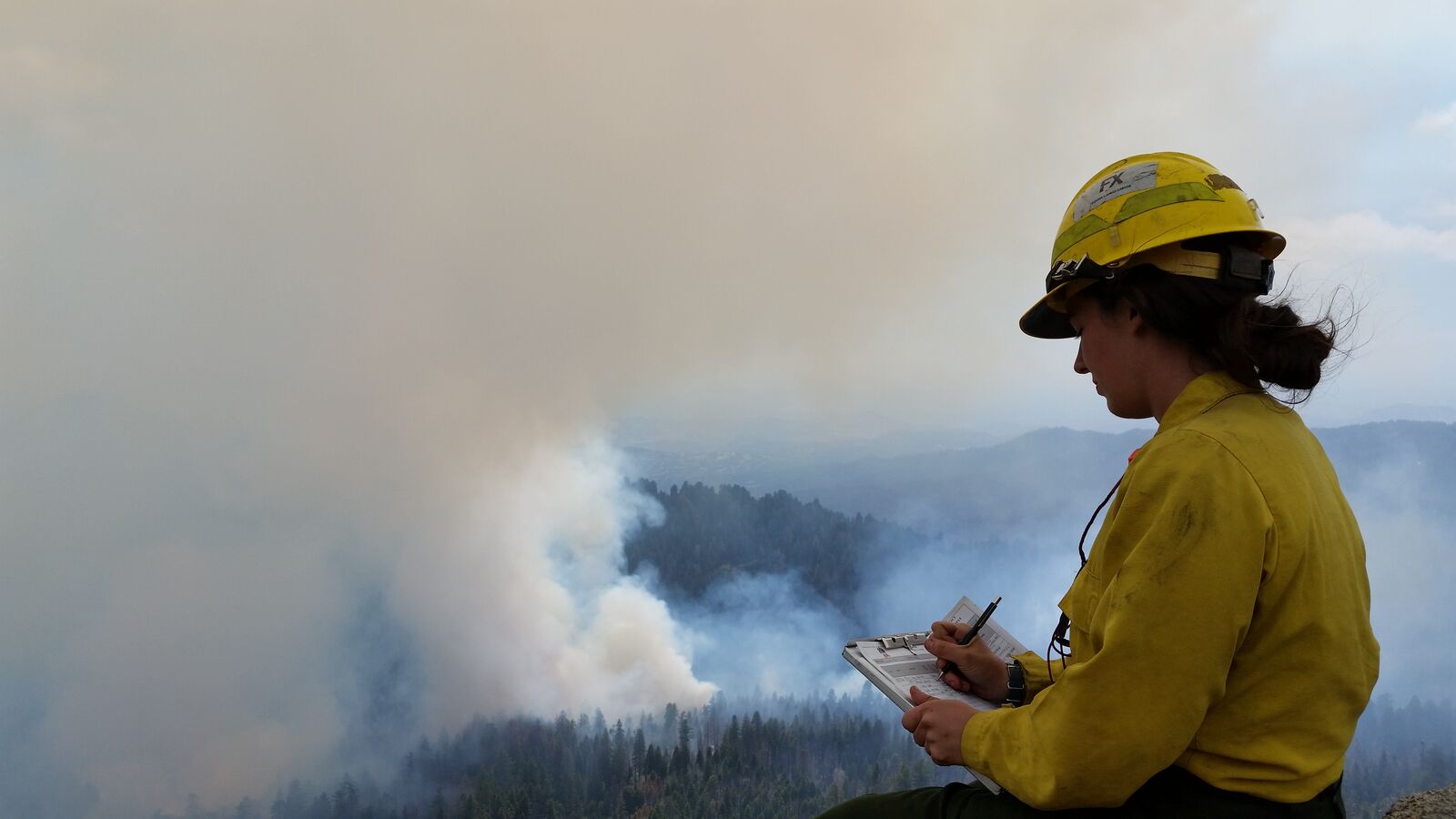 A fire effects monitor in a yellow Nomex shirt and yellow hard hat sits on a high ridge, writing on a clipboard with a pen. Below and in front of her, a broad column of white smoke rises out of forested ridges from a prescribed burn, filling most of the sky.
