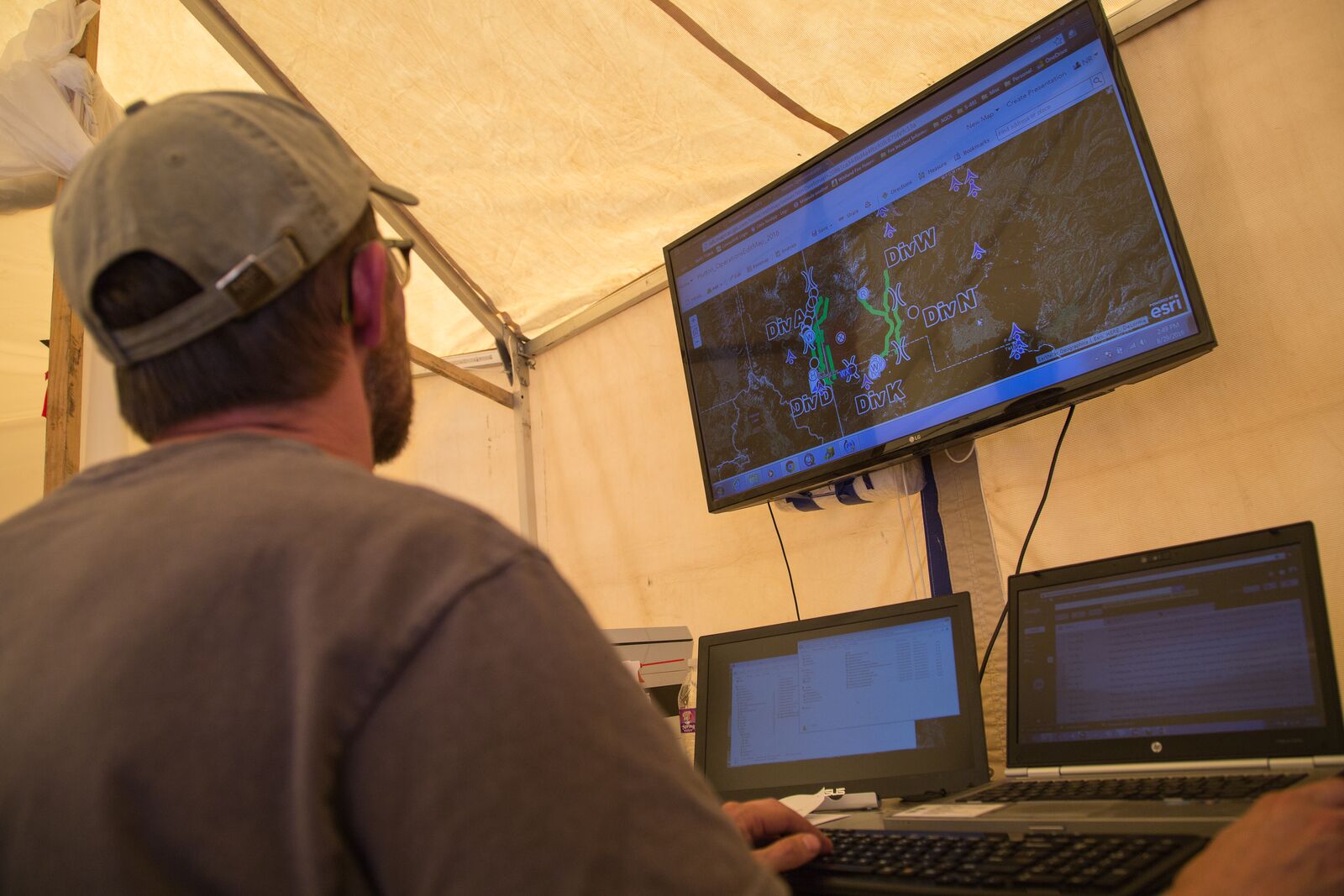 A GIS specialist in a grey ball cap, seen from behind, sits at a laptop inside a tent at an incident command post and looks up at a large screen mounted on the tent frame. The screen shows a mapping program: a dark terrain and satellite basemap overlaid with a bright fire perimeter line and labelled division breaks marking sections of the fire.