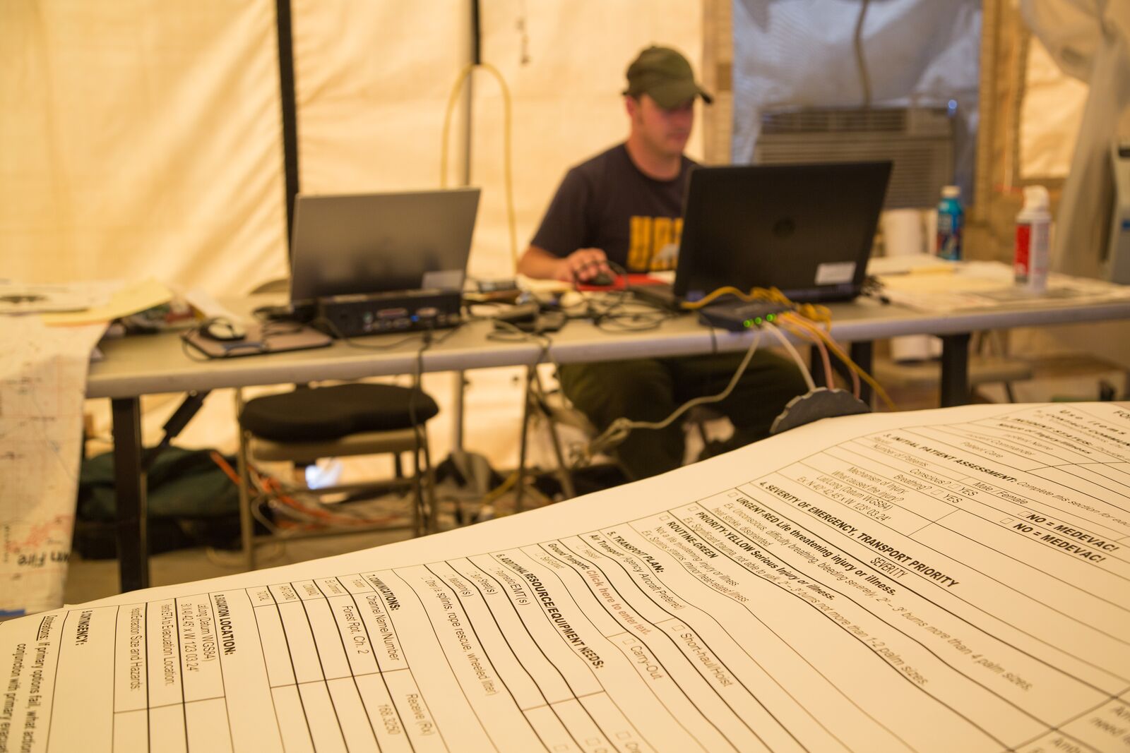 Inside the planning tent at a wildfire incident command post. A man in a ball cap sits at a folding table in a canvas wall tent, typing on one of two open laptops, with a power strip and bundles of orange and yellow network cables running across the table beside him. A printed incident command form, ruled into hand-labelled boxes, lies across the table in the foreground.