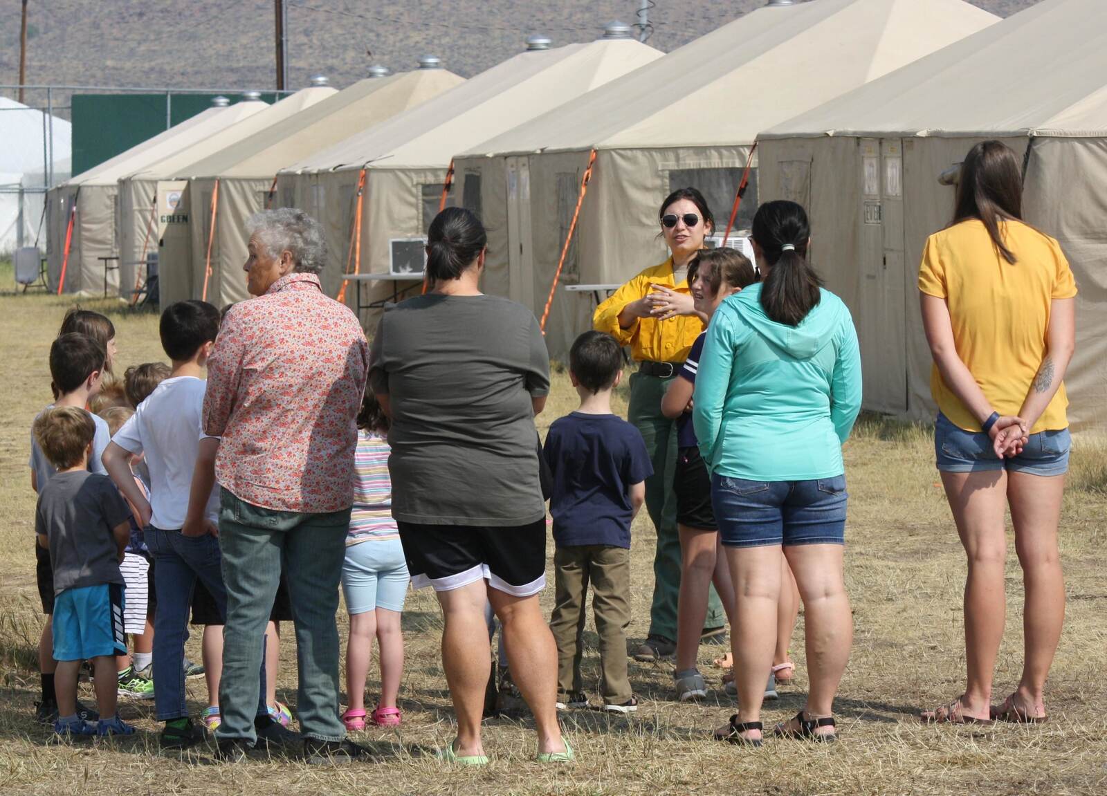 A public information officer in a yellow Nomex shirt and sunglasses stands in front of a long row of large canvas tents at a wildfire incident command post, talking and gesturing to a group of children and adults who have gathered on the dry grass in front of her to listen.